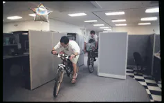 Two men ride bikes through a maze of cubicles. A silver balloon shaped like a star rises from one cubicle.