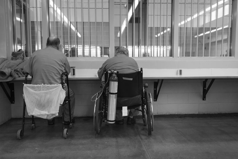 Two men in wheelchairs are photographed from behind in a prison setting; one uses an oxygen tank
