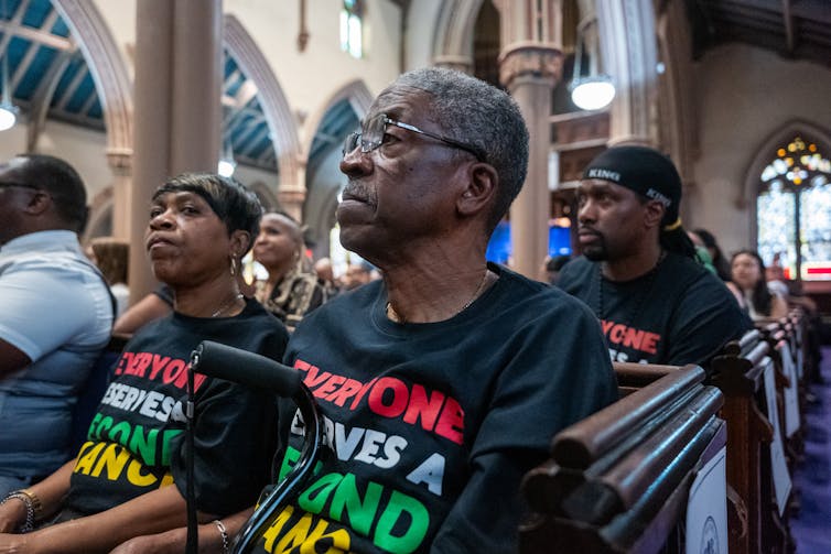 A group of mostly black men and women sat in a church, some wearing shirts that read
