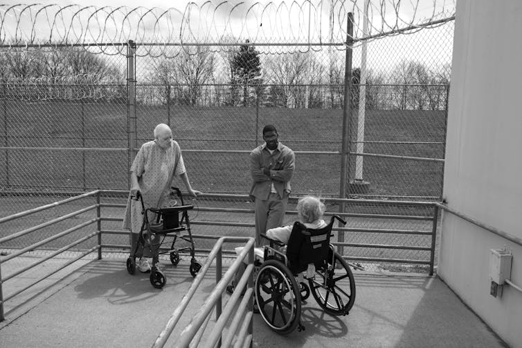 Three people stand outdoors in a prison yard; one uses a walker and another a wheelchair.