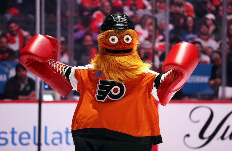 Large furry orange mascot with googly eyes wearing a sports jersey and boxing gloves
