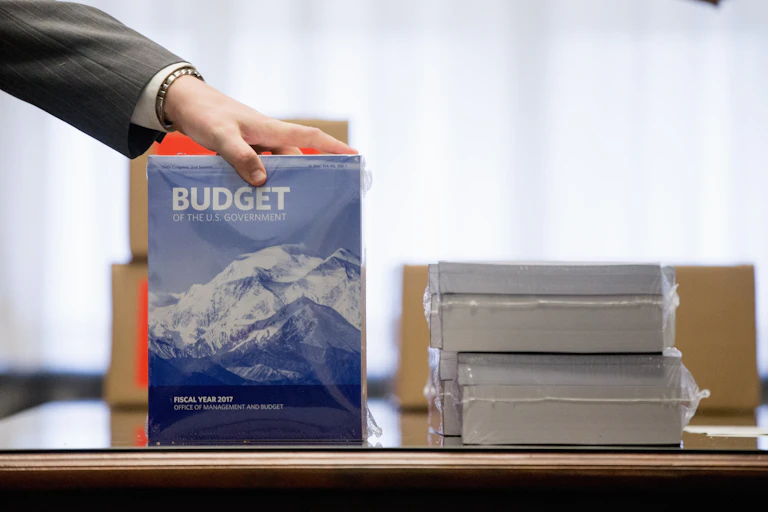 a hand holds a copy of the federal budget upright on a table next to a stack of big fat budget books