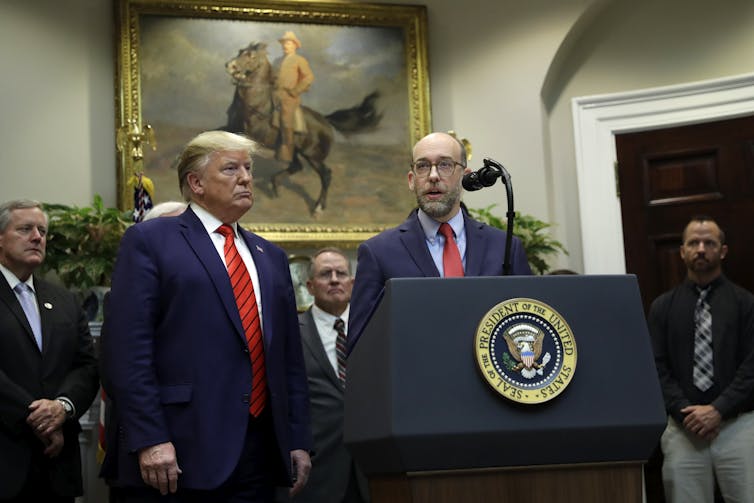 two men in suits and red ties stand in a room with other people and fancy paintings on the walls, while one stands in front of a podium with a mic