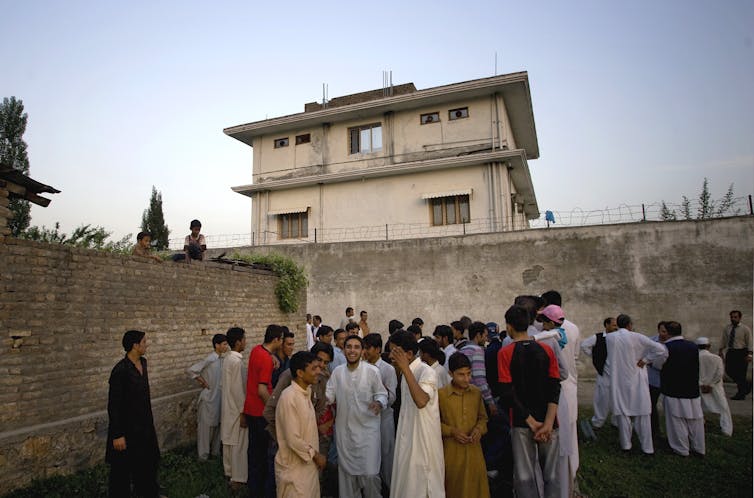 a group of people stand in front of a wall with a three-story concrete building in the background