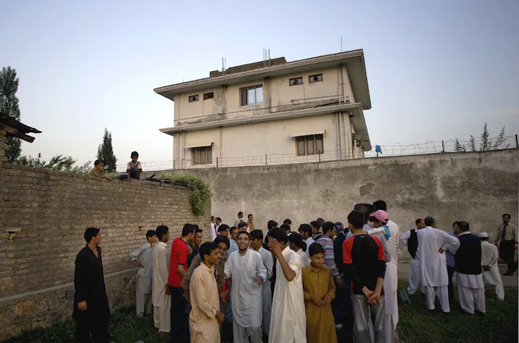 a group of people stand in front of a wall with a three-story concrete building in the background