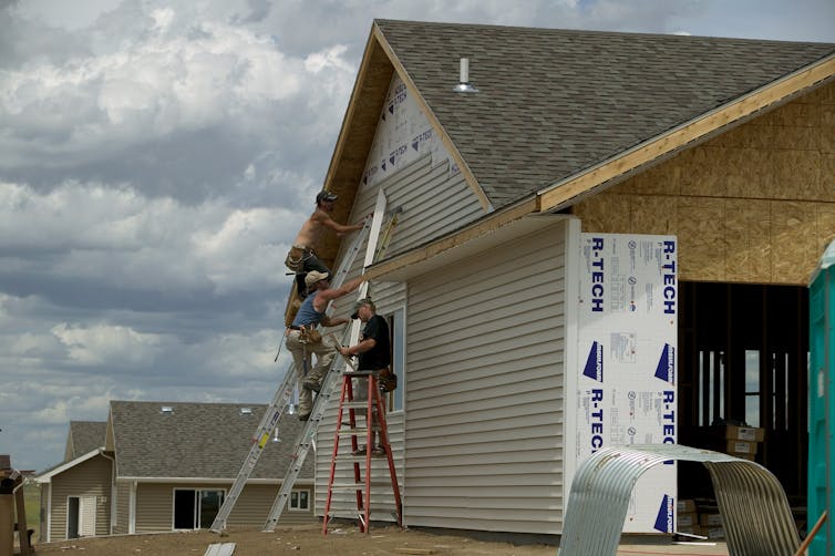 Construction workers work on a home in a new subdivision. One is on a ladder leaning against the outside of the house and the walls are unfinished.