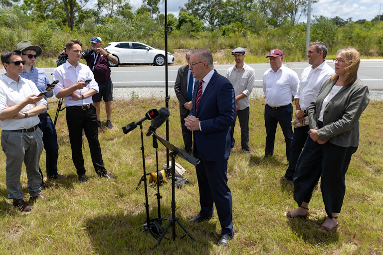 A man in a suit stands behind multiple microphones and speaks