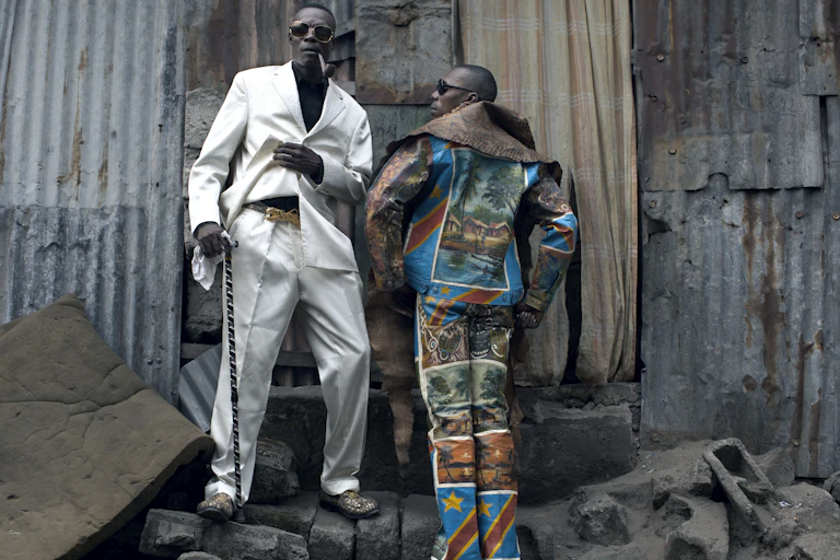 Two men pose in suits against the backdrop of a corrugated iron wall