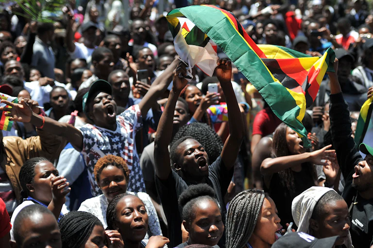A group of young people raise a Zimbabwean flag above their heads while shouting and waving their arms