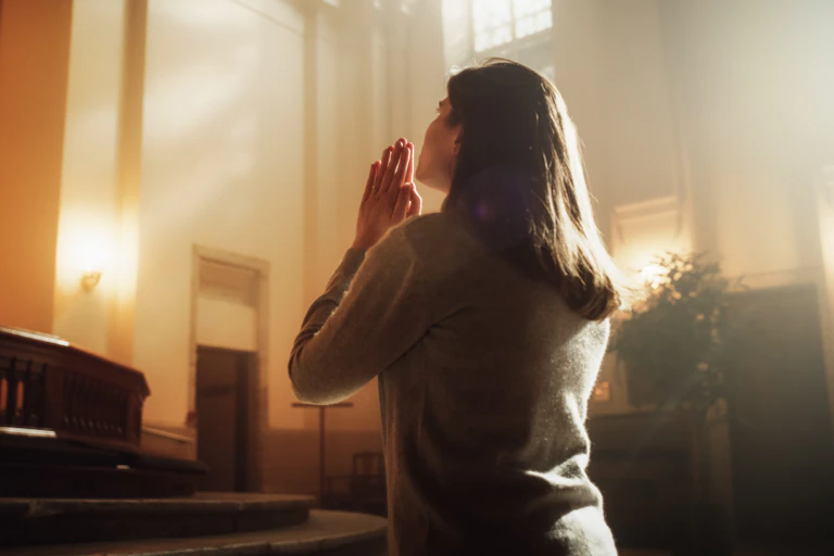 A woman in a church kneels and places her hands together in prayer.