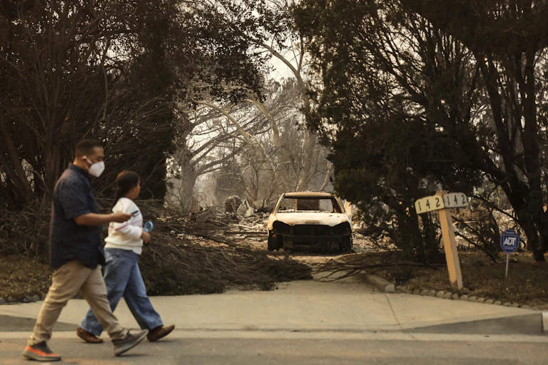 man and child walk past burned out car