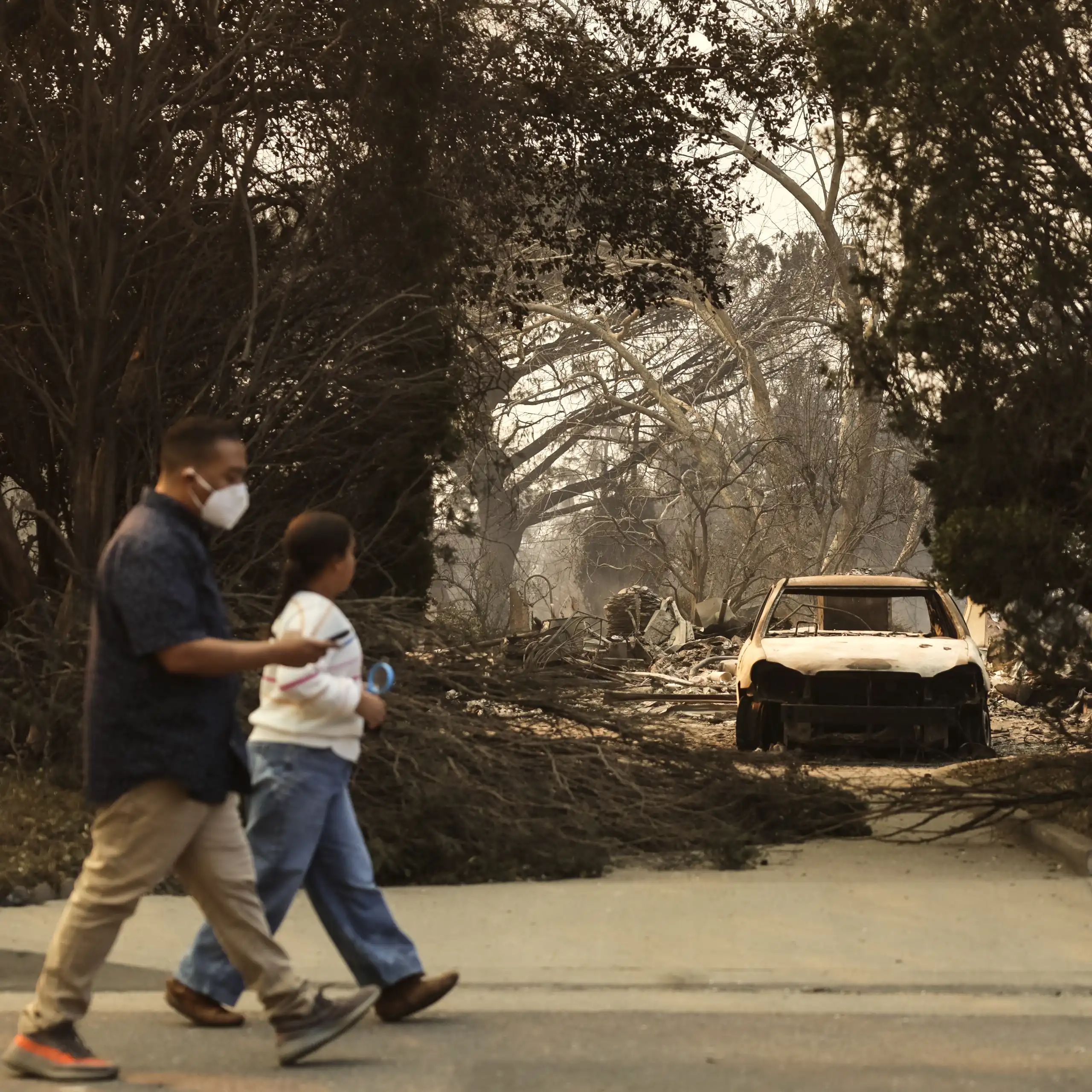 man and child walk past burned out car