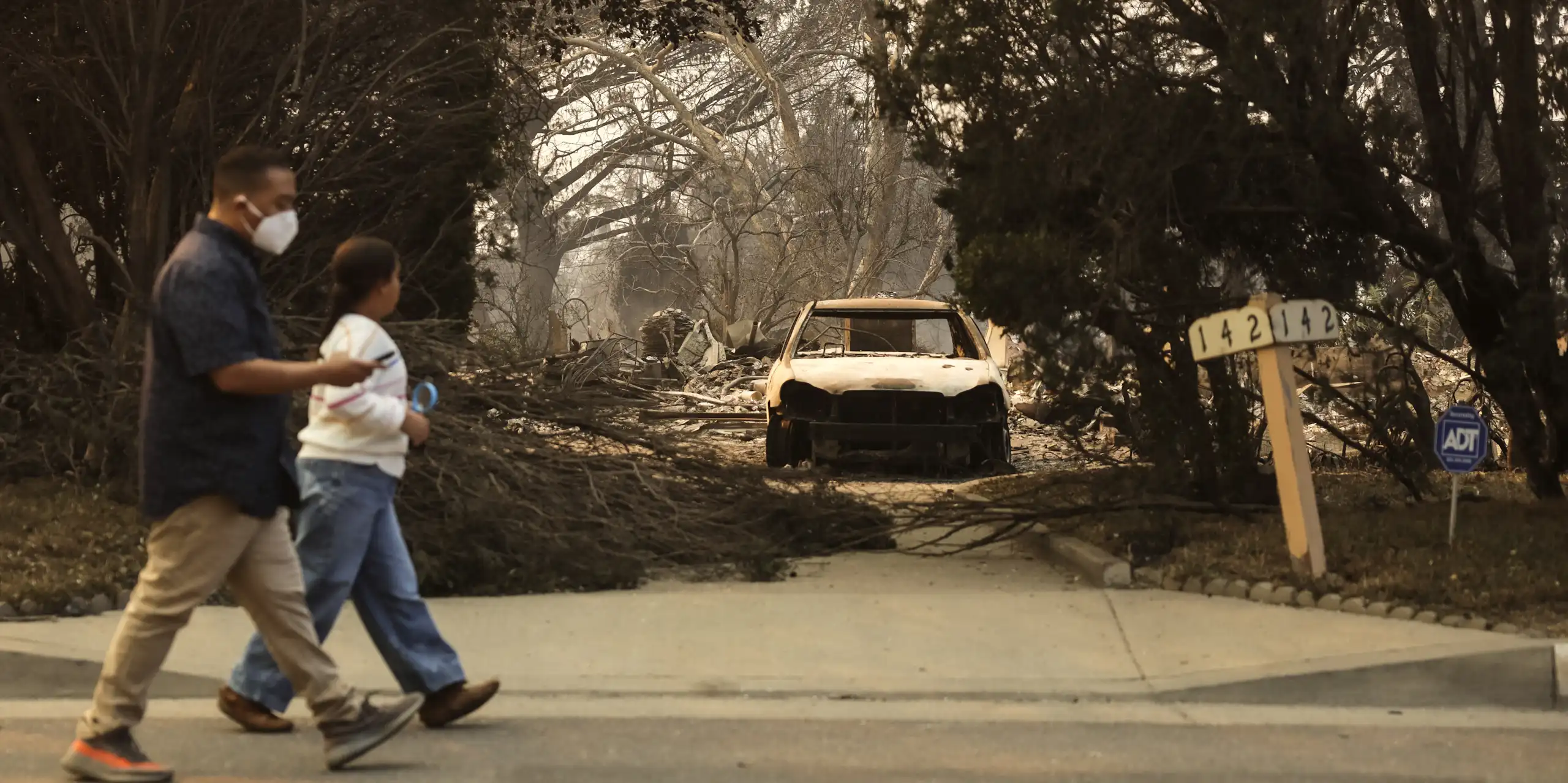man and child walk past burned out car