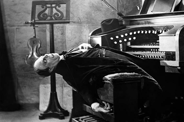 A demonic looking character in evening dress sitting leaning backwards at an organ.