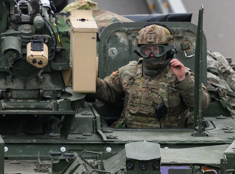 A person in a helmet and camouflage stands in an opening of a military vehicle.