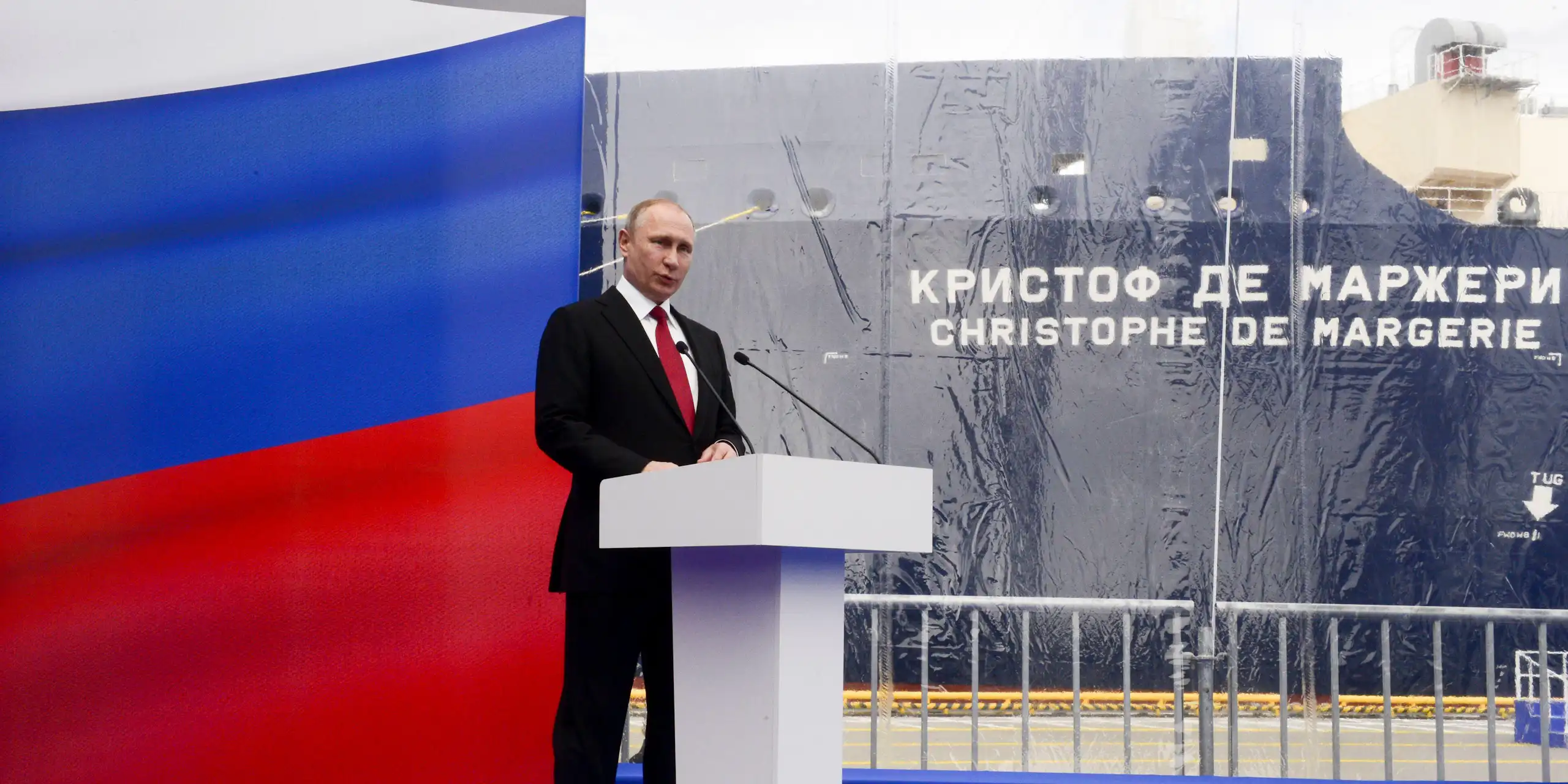 Vladimir PUtin at a podium with a Russian flag. In the background is the Christophe de Margerie LNG carrier.