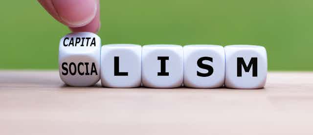A hand flips a dice -- one face of which says "CAPITA" and the other says "SOCIA" -- next to four more dice that together read "LISM."