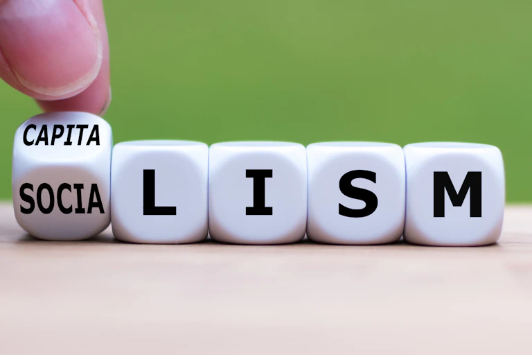 A hand flips a dice -- one face of which says "CAPITA" and the other says "SOCIA" -- next to four more dice that together read "LISM."