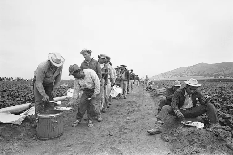 Farmworkers line up for lunch in a field in an old black and white photo.