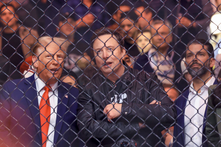 Donald Trump, Elon Musk and Donald Trump Jr. watch something behind a fence amid a big crowd.