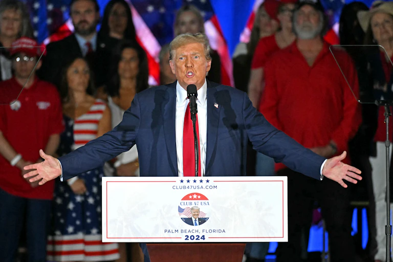 Donald Trump stands behind a lectern, his arms outstretched.