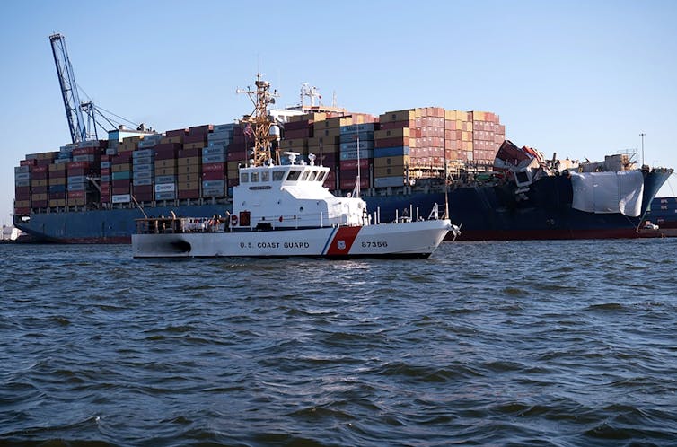 A white ship with an orange stripe sails in front of a large container ship.