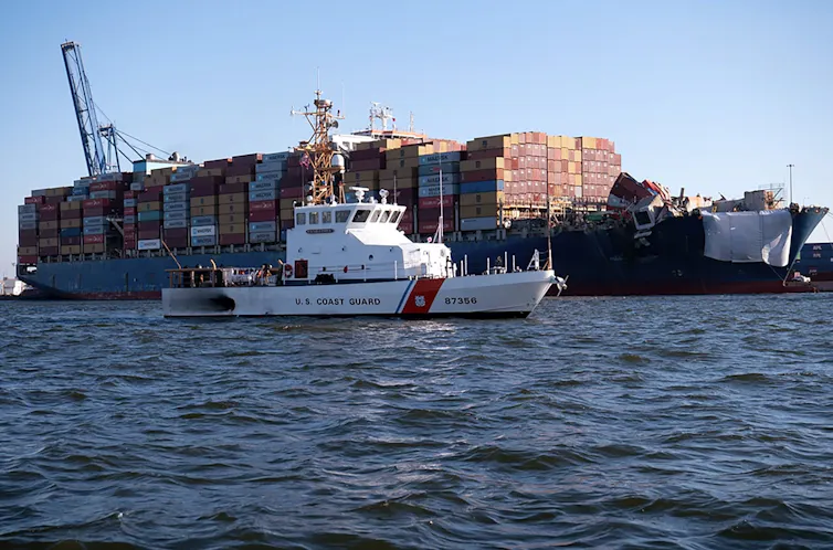 A white ship with an orange stripe sails in front of a large container ship.
