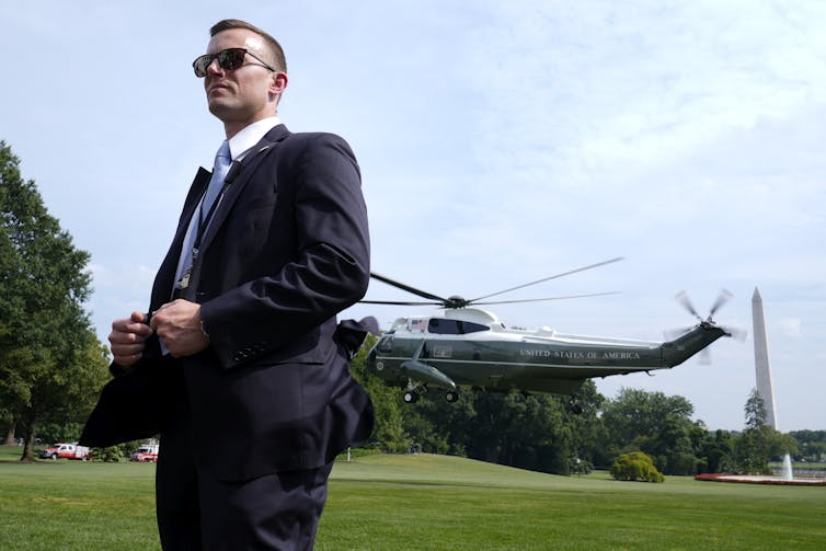 A person in a suit and sunglasses stands outside in front of a Marine helicopter.
