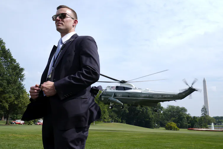 A person in a suit and sunglasses stands outside in front of a Marine helicopter.