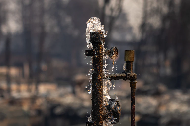 Water bubbles up out of a pipe surrounded by burned landscape. Only the foundation of the house remains.
