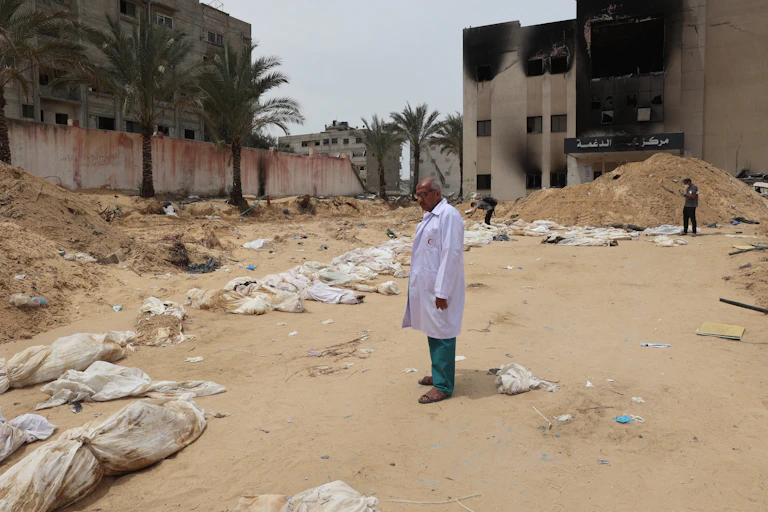 A man in a white doctors jacket stands in front of body bags.