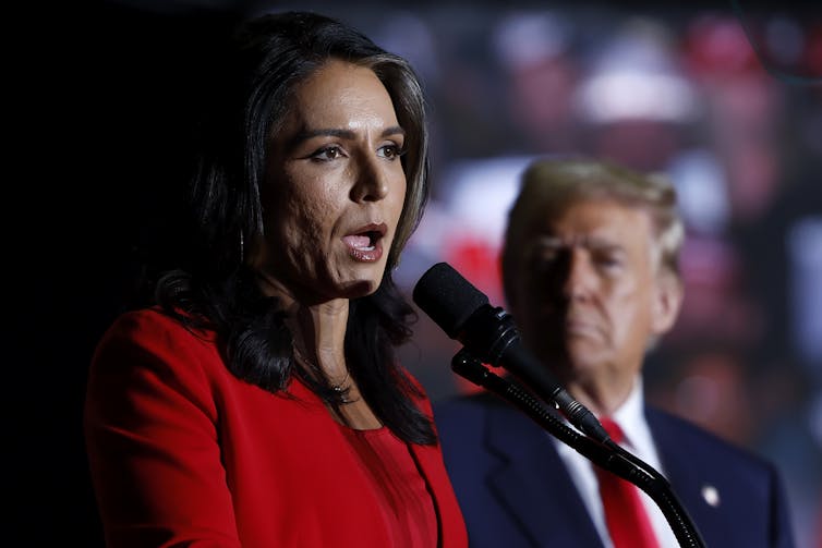 A dark-haired woman in a red top speaks into a microphone with a man in a suit and tie next to her.