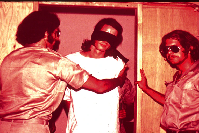 Two men in fake guard uniforms handle a man in a white shirt and blindfold.