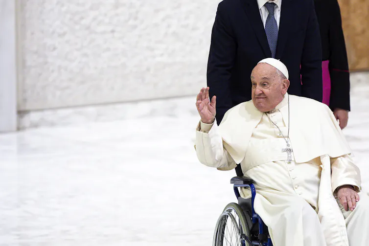 Pope Francis, dressed in white and in a wheelchair, waves to someone in the distance.