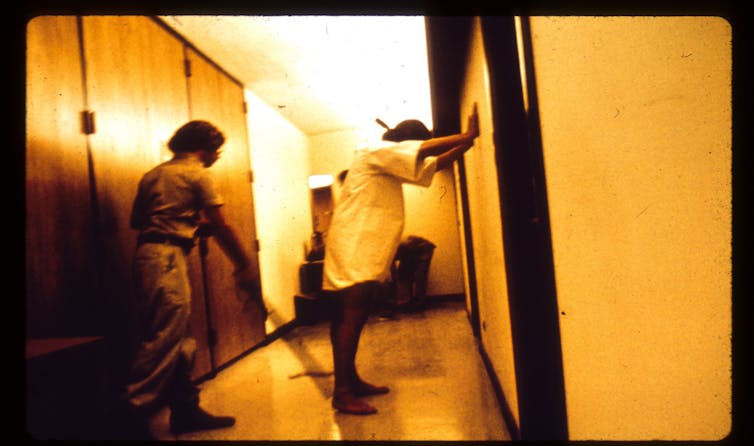 A man in a short white shirt stands with his hands on the wall while a man in a uniform directs him with a police baton.