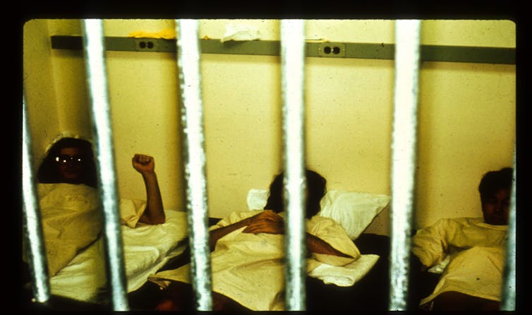 An old photo of three figures lying in beds with bars visible in the foreground.