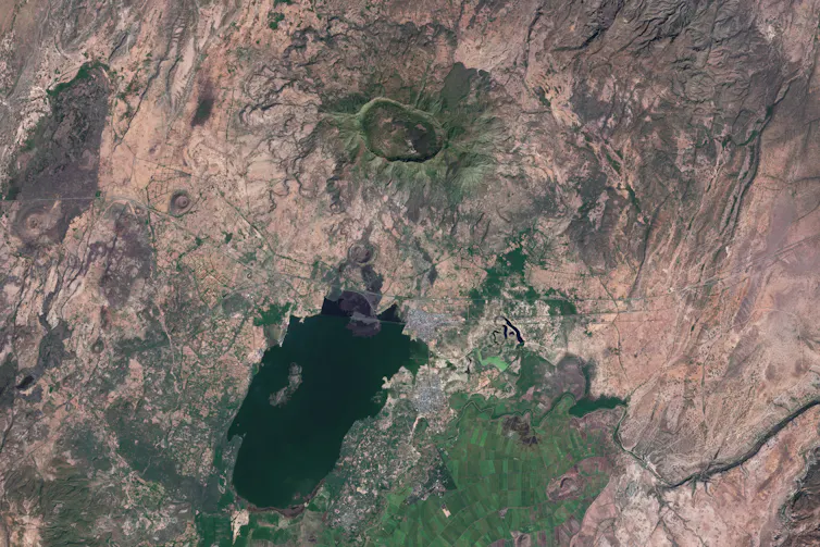 A satellite image that shows a big depression amid a brown and green landscape. There is also a large body of water visible