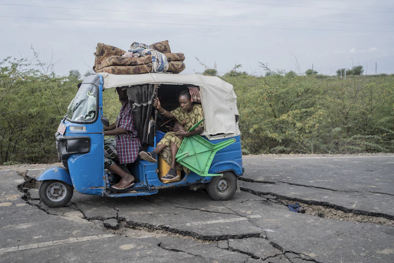 A woman lifts her feet as the tiny blue and white vehicle she's traveling in navigates wide fissures in the road