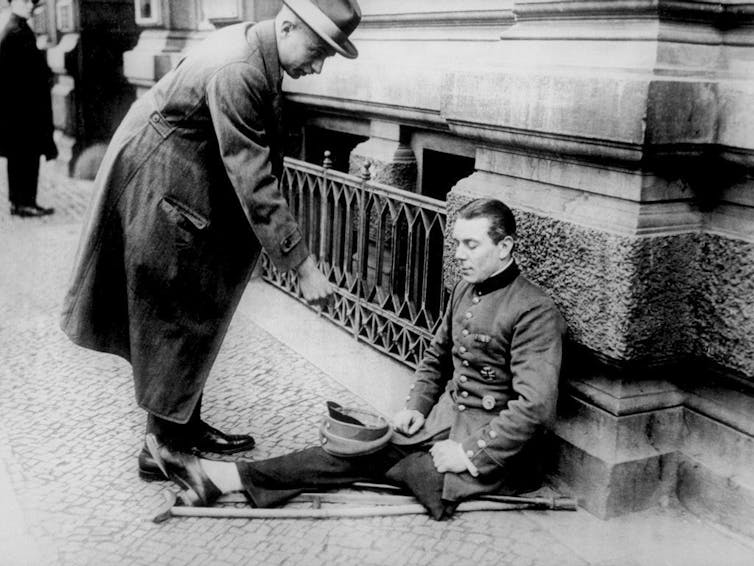 Un hombre da limosna a un antiguo soldado que ha perdido una pierna y está sentado en la acera.
