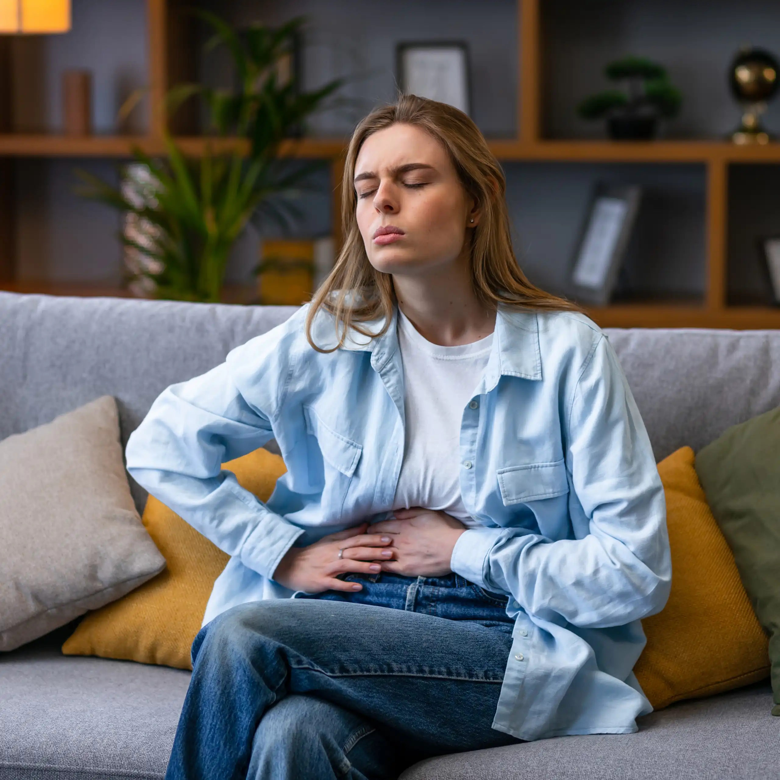 A young woman sitting on a couch holds her stomach in pain.