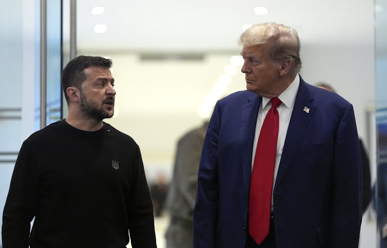 A rotund man in a blue suit and red tie with fluffy greyish blond hair talks to a shorter dark-haired man in a black shirt.