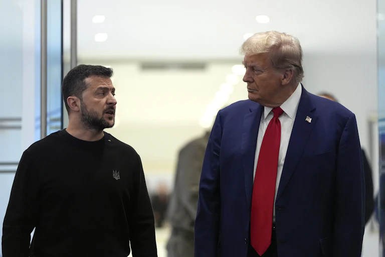 A rotund man in a blue suit and red tie with fluffy greyish blond hair talks to a shorter dark-haired man in a black shirt.