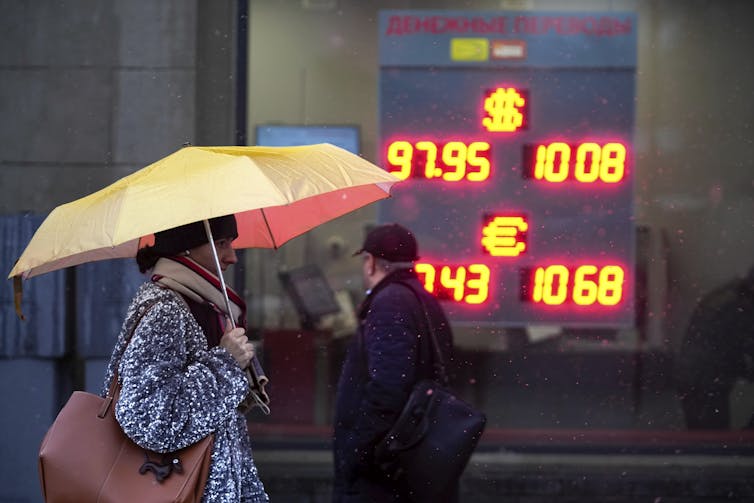 A woman carrying an umbrella walks past a sign lit up with numbers.