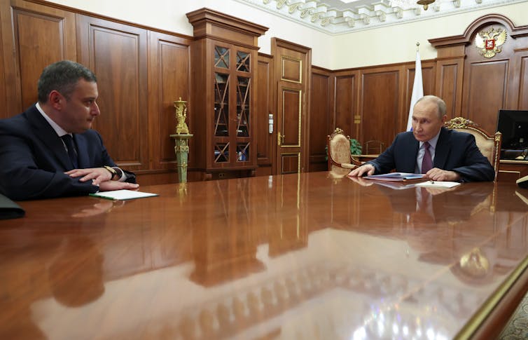 Two men sit far apart at a gleaming wooden table.