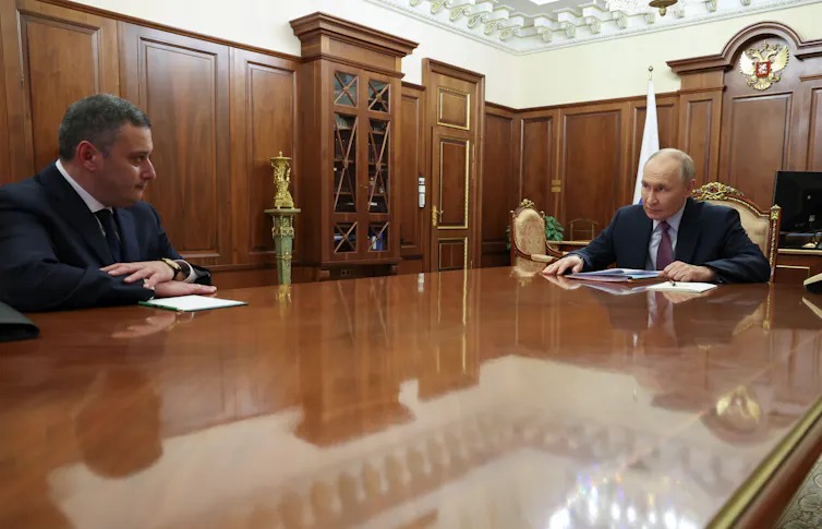 Two men sit far apart at a gleaming wooden table.