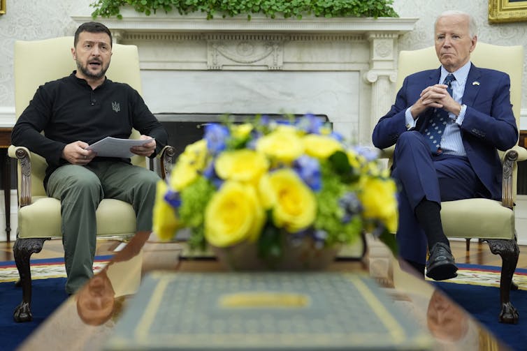 A dark-haired bearded man sits in an ornate office next to an older man with grey hair.
