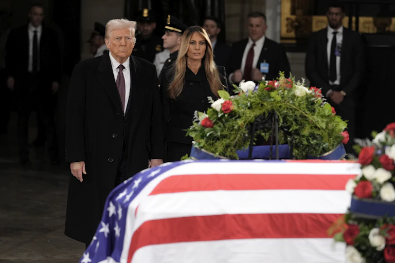 A man with fluffy grey-blond hair stands next to a woman with long dark-blond hair behind a flag-draped casket.