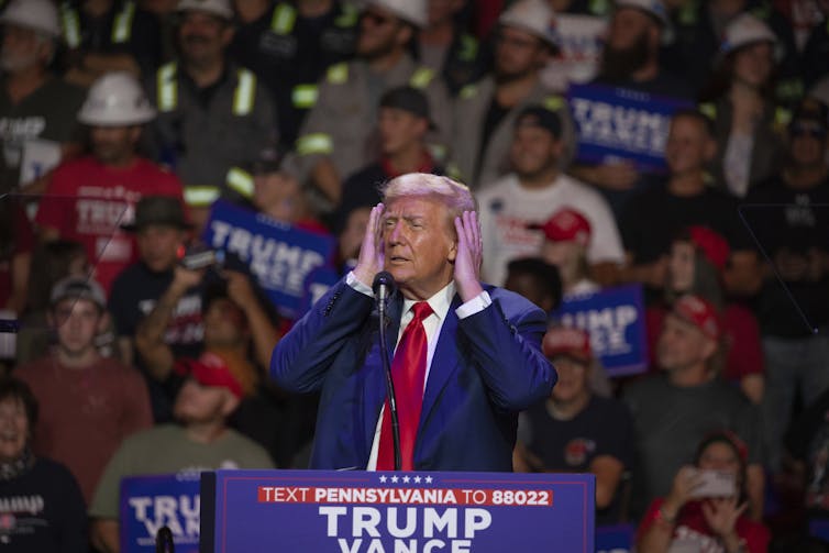 A man in a blue suit and red tie stands on a stage checking out his fluffy grey-blond hair on a large screen.