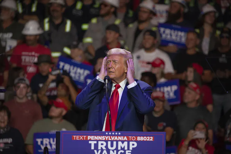 A man in a blue suit and red tie stands on a stage checking out his fluffy grey-blond hair on a large screen.