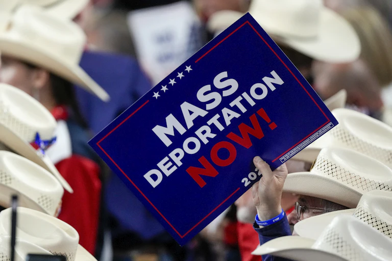 A person holding a sign reading "MASS DEPORTATION NOW!" among rows of people wearing white cowboy hats
